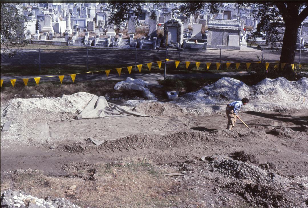 A man with a shovel in a construction zone. Yellow flags separate the sidewalk from the road under construction; above ground tombs from Greenwood Cemetery are in the background.