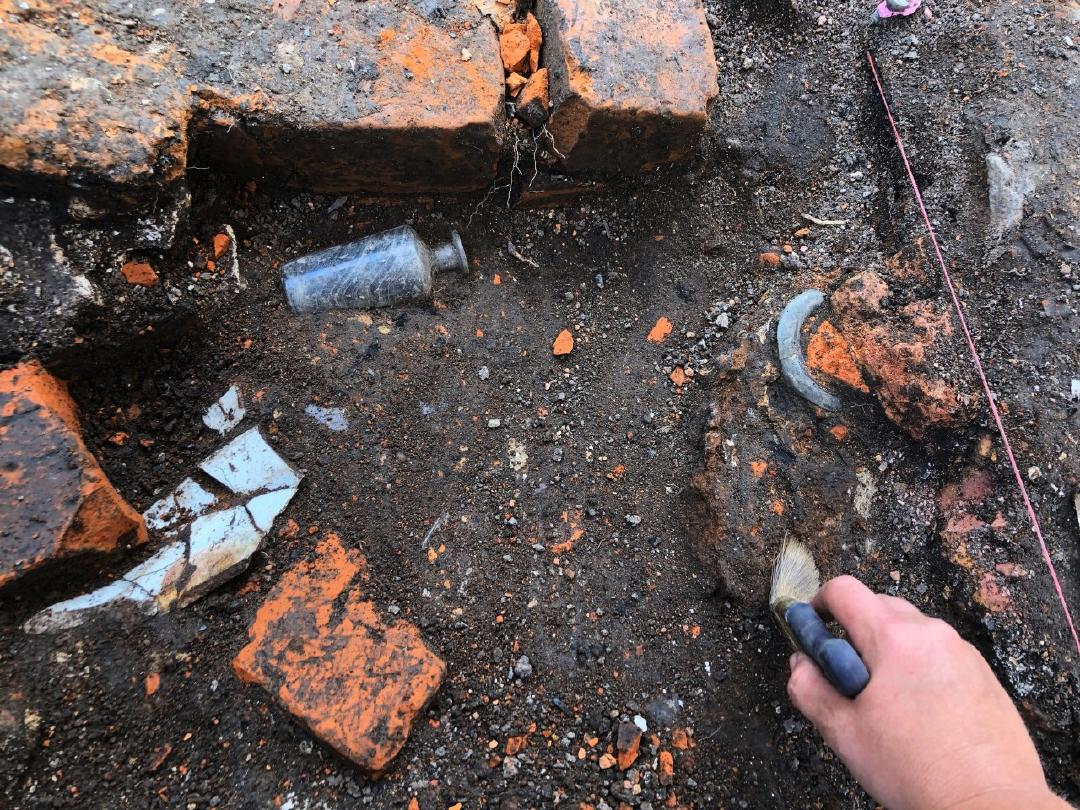 Showing brick, a bottle and ceramics embedded in soil and a hand brushing dirt off of a brick<br />
<br />