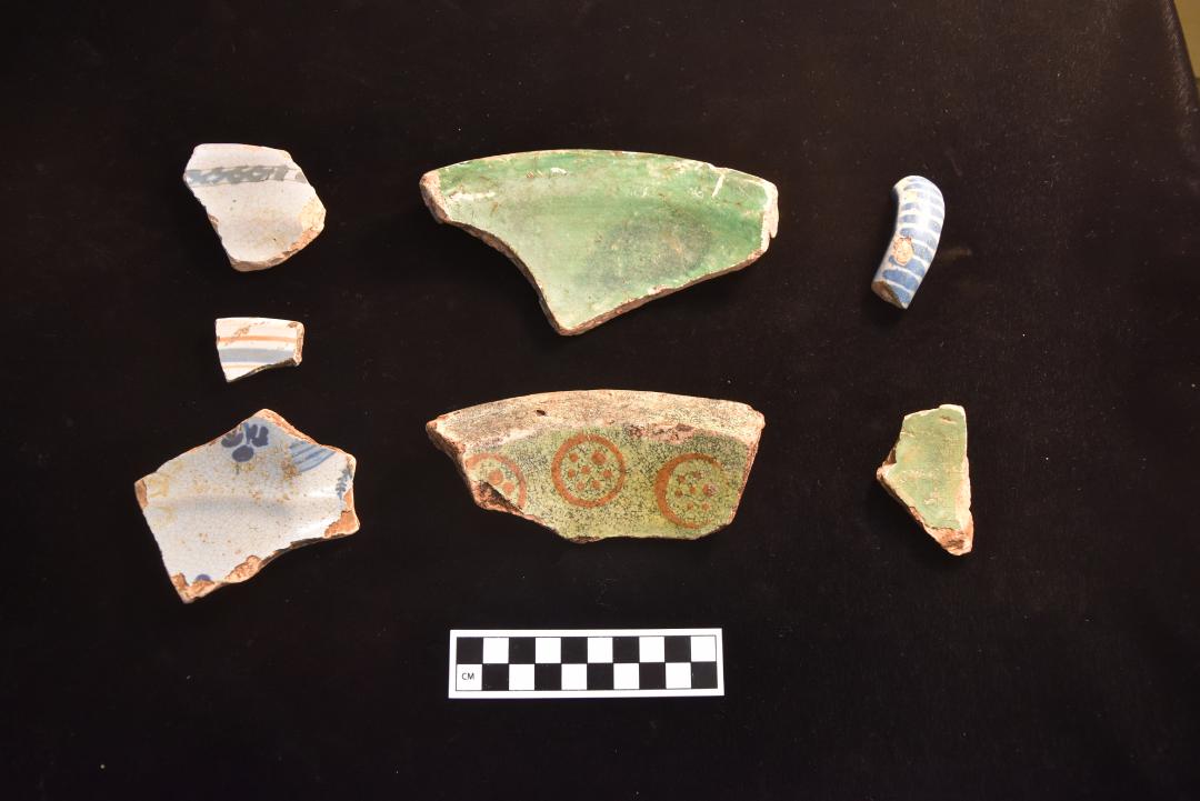 Photograph of ceramic pottery fragments. The pottery is arranged in two neat rows on a black back drop with a white and black scale bar displaying centimeters. The top row shows three ceramic types. From left to right: a sherd of a plate rim with a blue painted line, the second, a green rim fragment of a bowl/milk pan, and the third is a handle of a cup painted with white and blue stripes. The second row is as follows. Left to right: a white pottery fragment with blue dots and lines. The second, a yellow bowl or basin rim with orange circle and dot decoration, and the third a lime green pottery fragment.