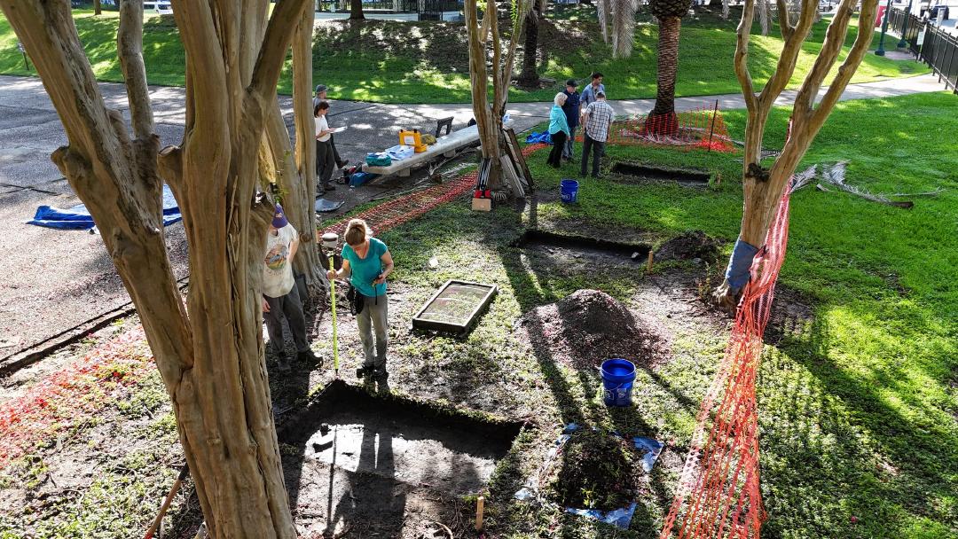 Drone shot from above of clusters of people outside, under trees, around 1 x 2 meter units. In the foreground, two people are standing with survey equipment.