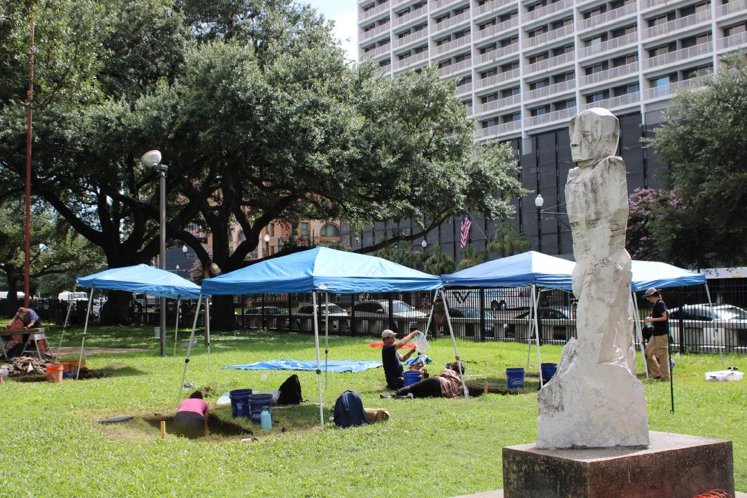Overview photograph of the Duncan Plaza Archaeological Field School, showing an active archaeological excavation in an open grassy plaza with oak trees and a statue.