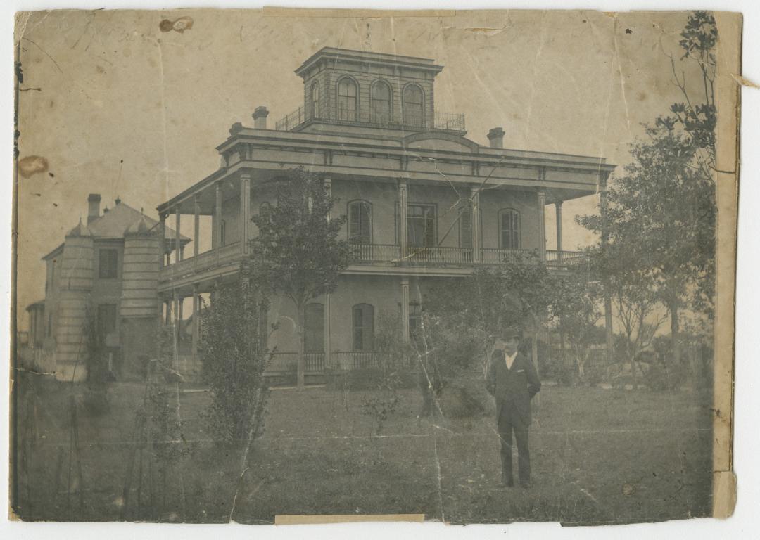 Historic sepia-toned photograph showing a man in suit and bowler standing in front of a large home with two-story wraparound porch and topped with a cupola. All window openings are large, shuttered and arched. To the left are two round towers and another, slightly smaller building in the background.