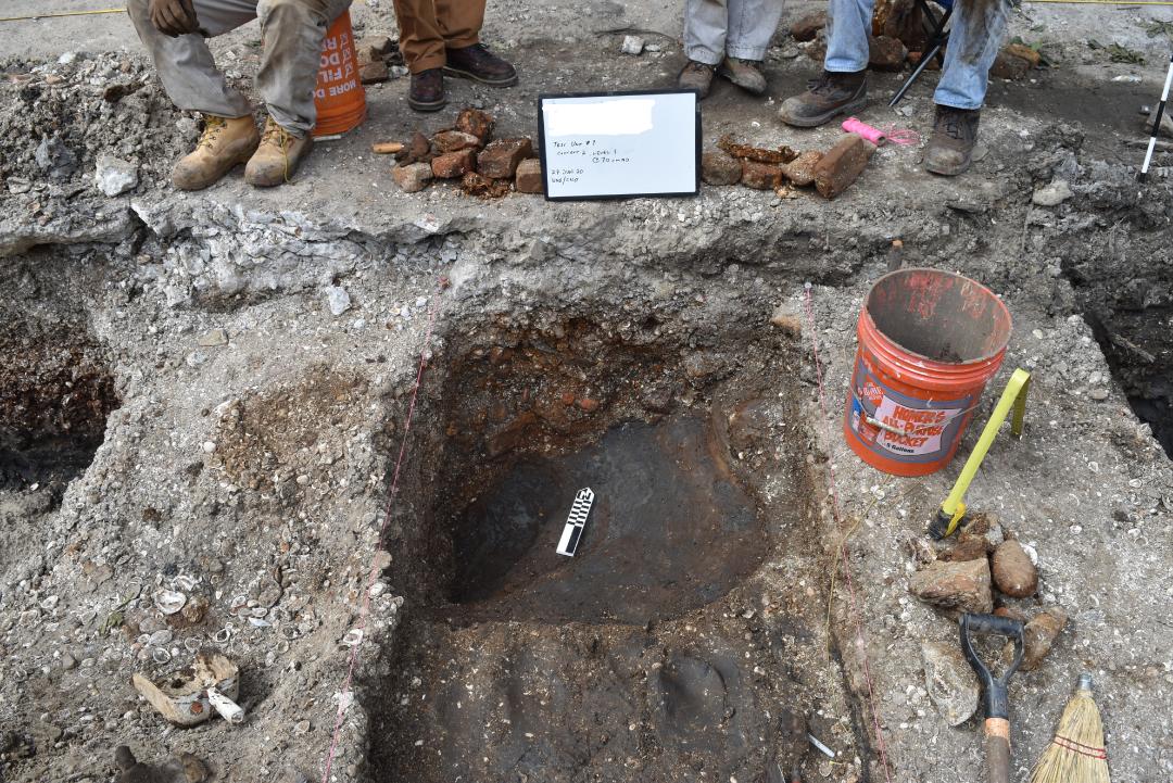 The photograph centers on a rectangular pit in the ground, flanked by a bright orange bucket and a white sign that reads, "Test Unit 1, Context 2, Level 1, @ 70 cmbd, 27 July 2020, UNO and CNO". A black and white arrow is in the middle of the pit indicating north. The photographer frames four pairs of feet standing on the ground surface above the pit in the background of the photograph.