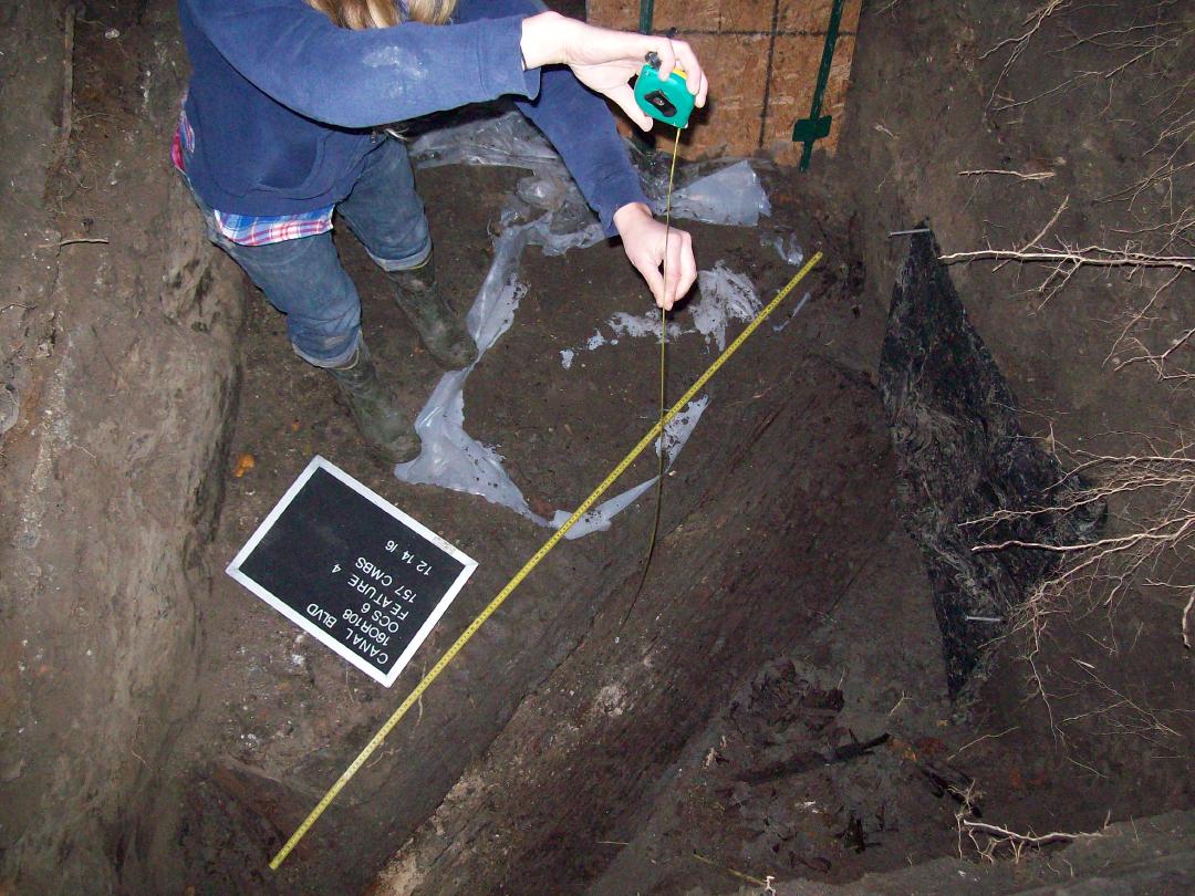 Person, whose face is not shown, is photographed in a pit, is holding a tape measure up and down next to a yellow tape measure along the side of a wooden coffin lid.