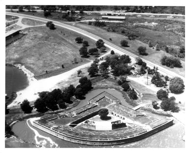 Black and white aerial view of a quarter circle shaped fort and the surrounding area with roads and trees.