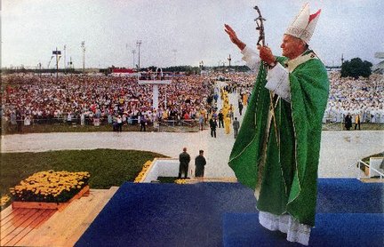 The pope, wearing a green robe, stands on a blue platform with arms raised while a dense crowd of onlookers are crowded in the background.
