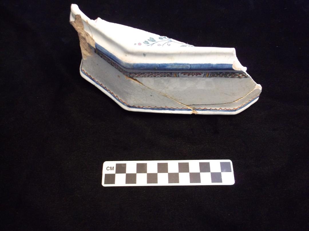 A ceramic lid fragment, white with blue edges and red, yellow, and green in-lined decoration; a flower or vine decorates the top of the fragment.