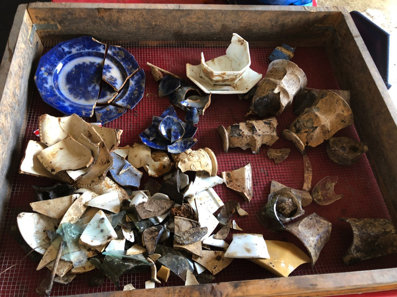 A square wood framed screen with wire mesh at the bottom containing broken historic ceramics and glass