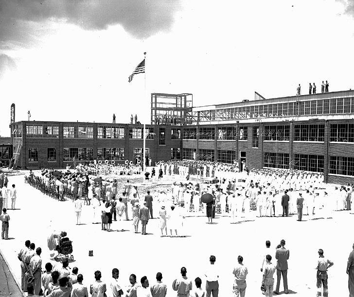 Black and white photo of a large group of people facing an American flag on a central flag pole in front of a partially built building. A large brass band plays in the background.