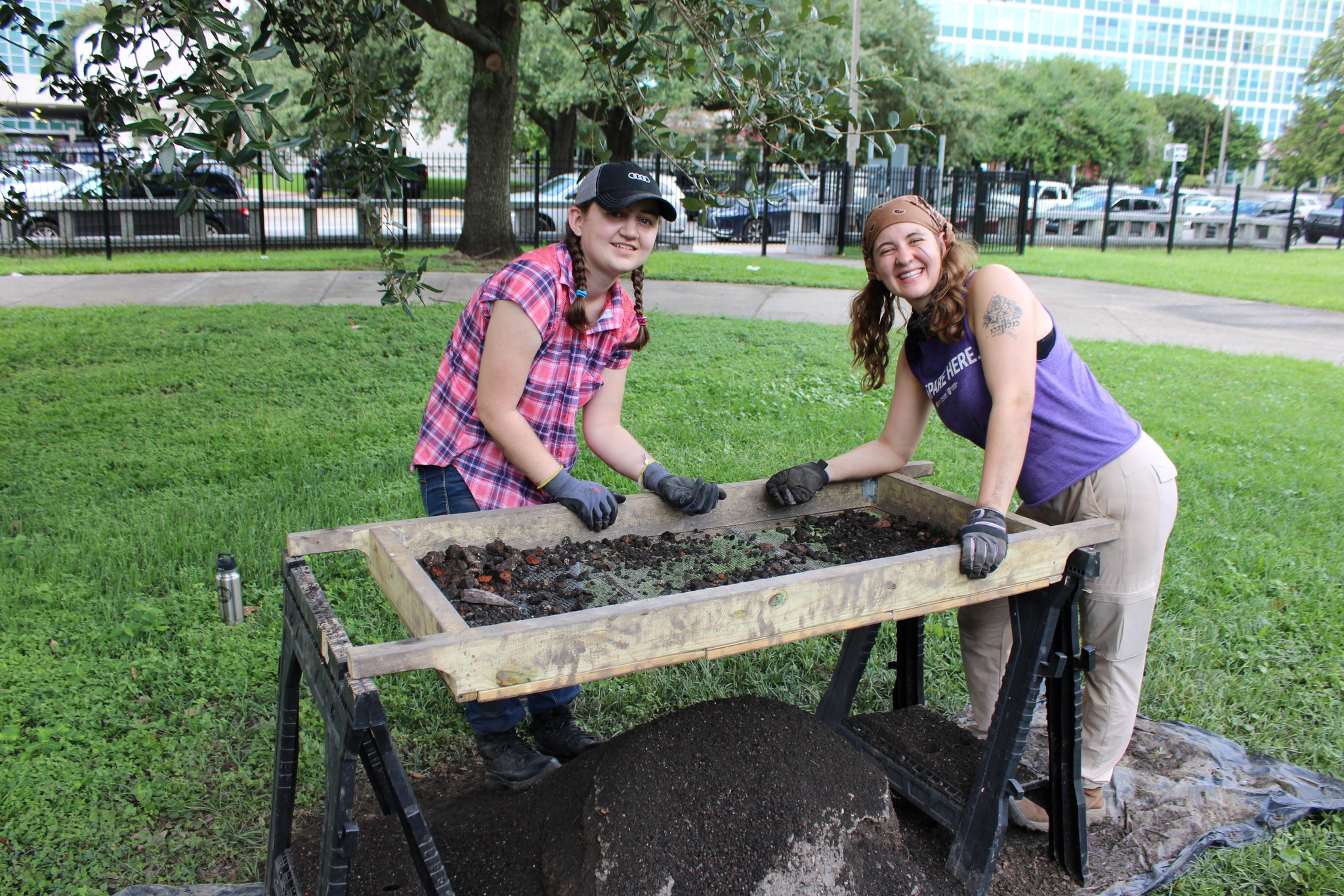 Photograph of two students around a wooden rectangular screen frame propped up on saw horses. A mound of dirt has been sifted through the quarter inch mesh screen.