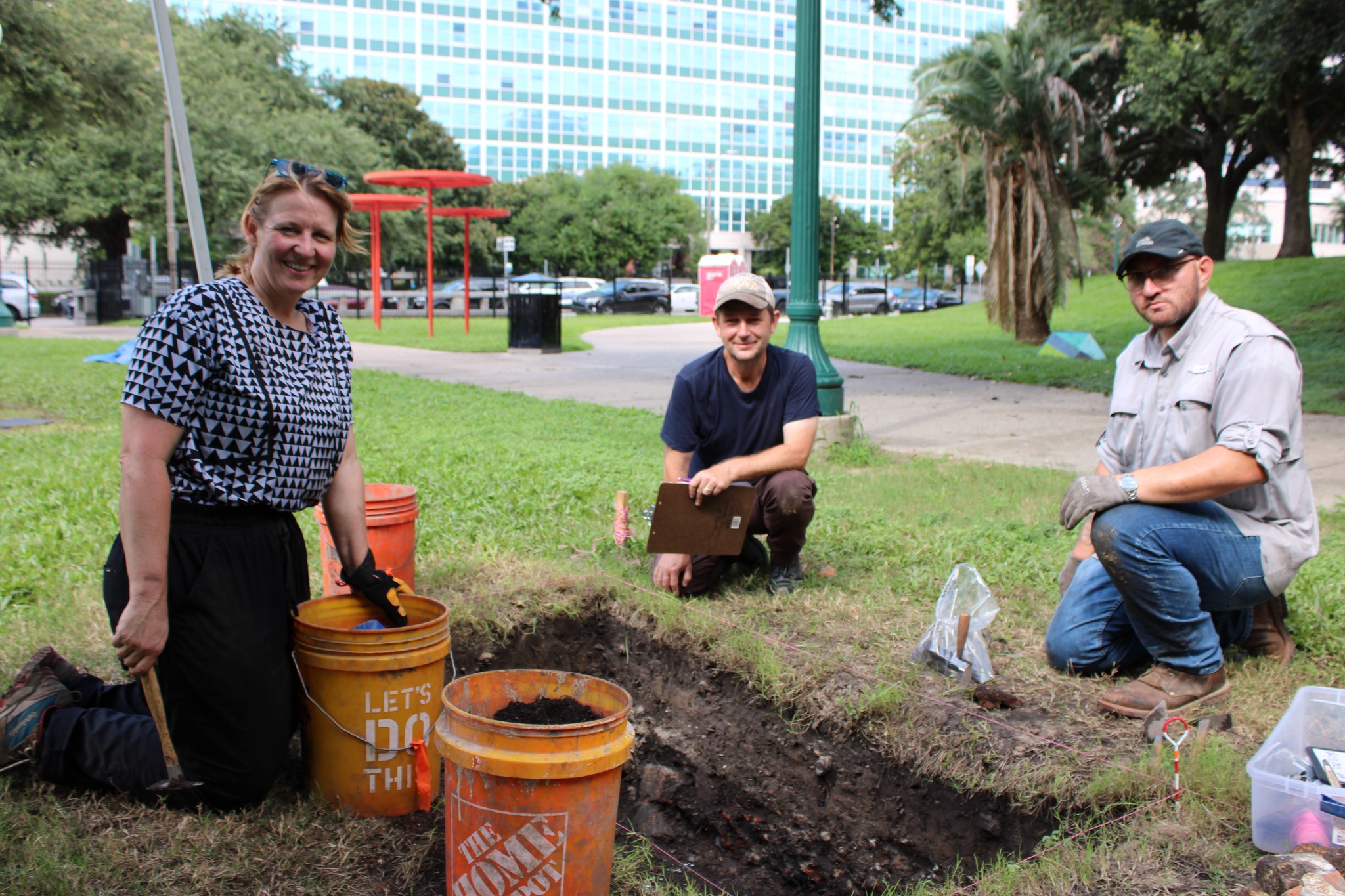 Photograph of three archaeologists smiling at the camera. In between them is a deep, rectangular excavation unit, orange buckets, and tools.