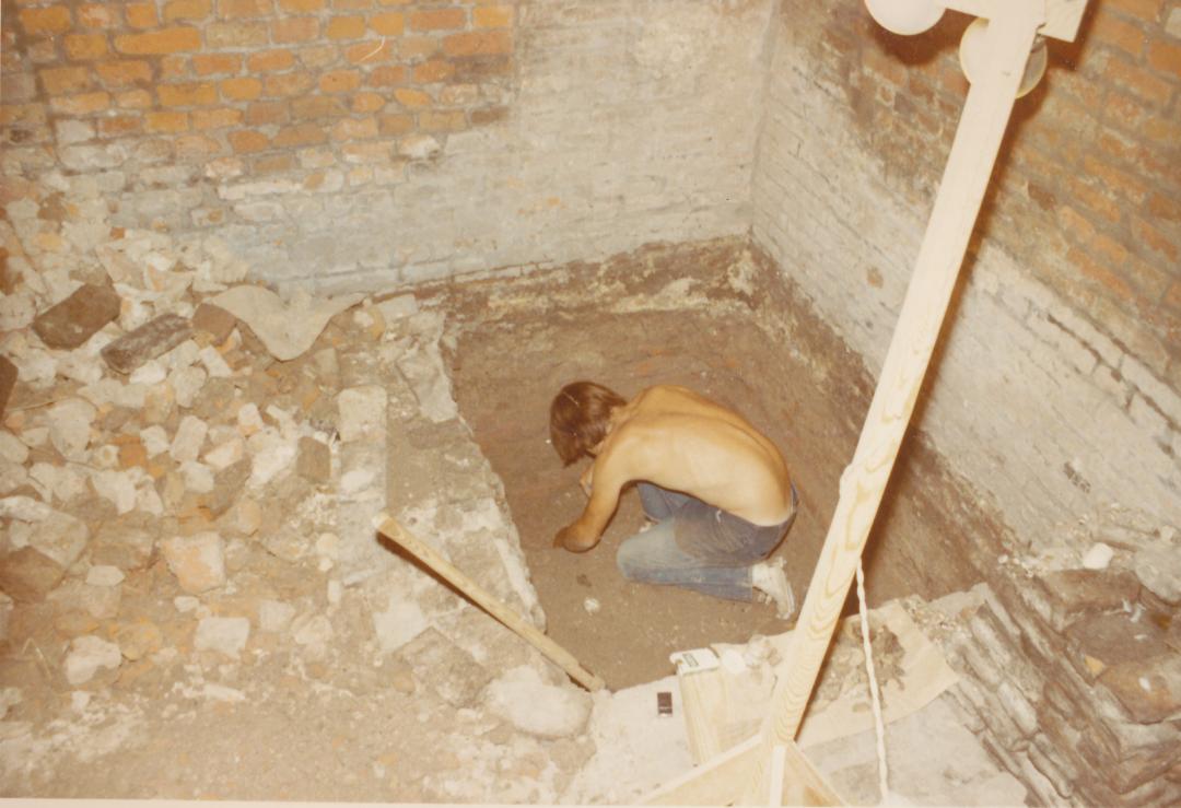 A young man excavates in a square pit in the corner formed by two brick walls. Brick rubble is on the ground surface to his left.