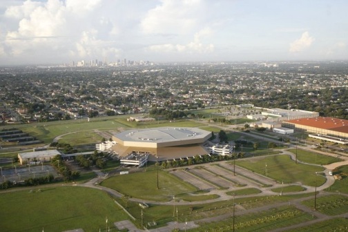 Aerial view of a large geometric building with a flat top situated within a green space.