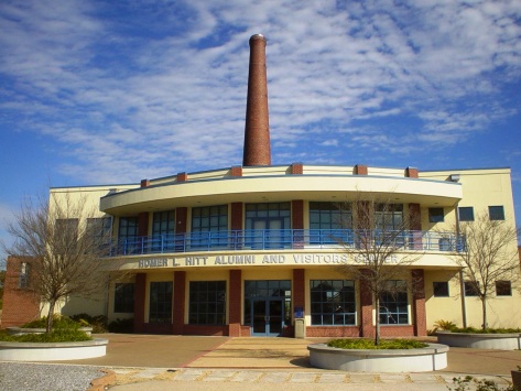 Brick and tan curved building facade with large brick smoke stack rising up behind it.